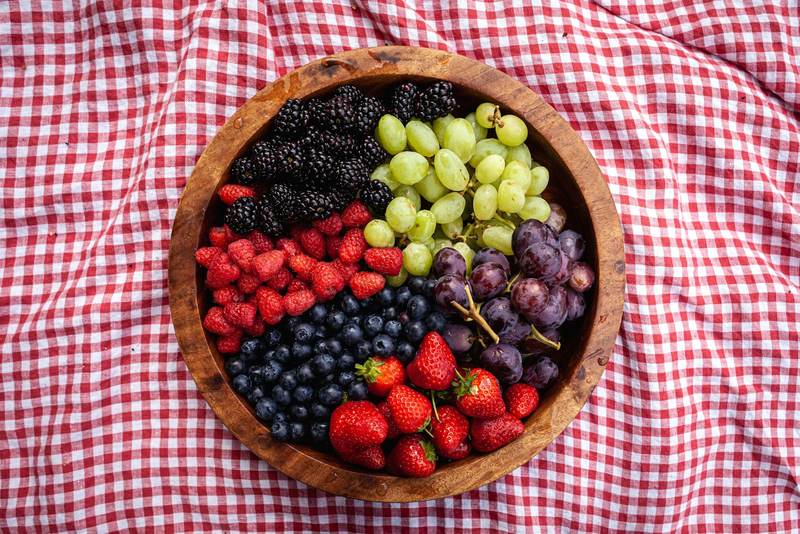 A wooden bowl of raspberries, blueberries, grapes and strawberries on a checked cloth