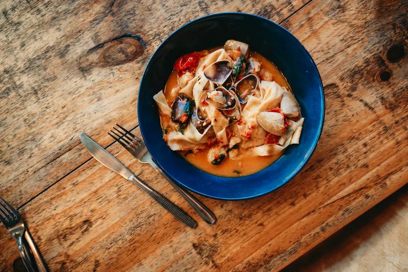 A blue bowl of seafood pasta on a wooden table with a knife and fork